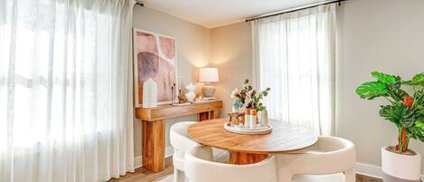 Un comedor con una mesa de madera y sillas blancas, una lámpara, una planta en maceta y un cuadro en la pared.