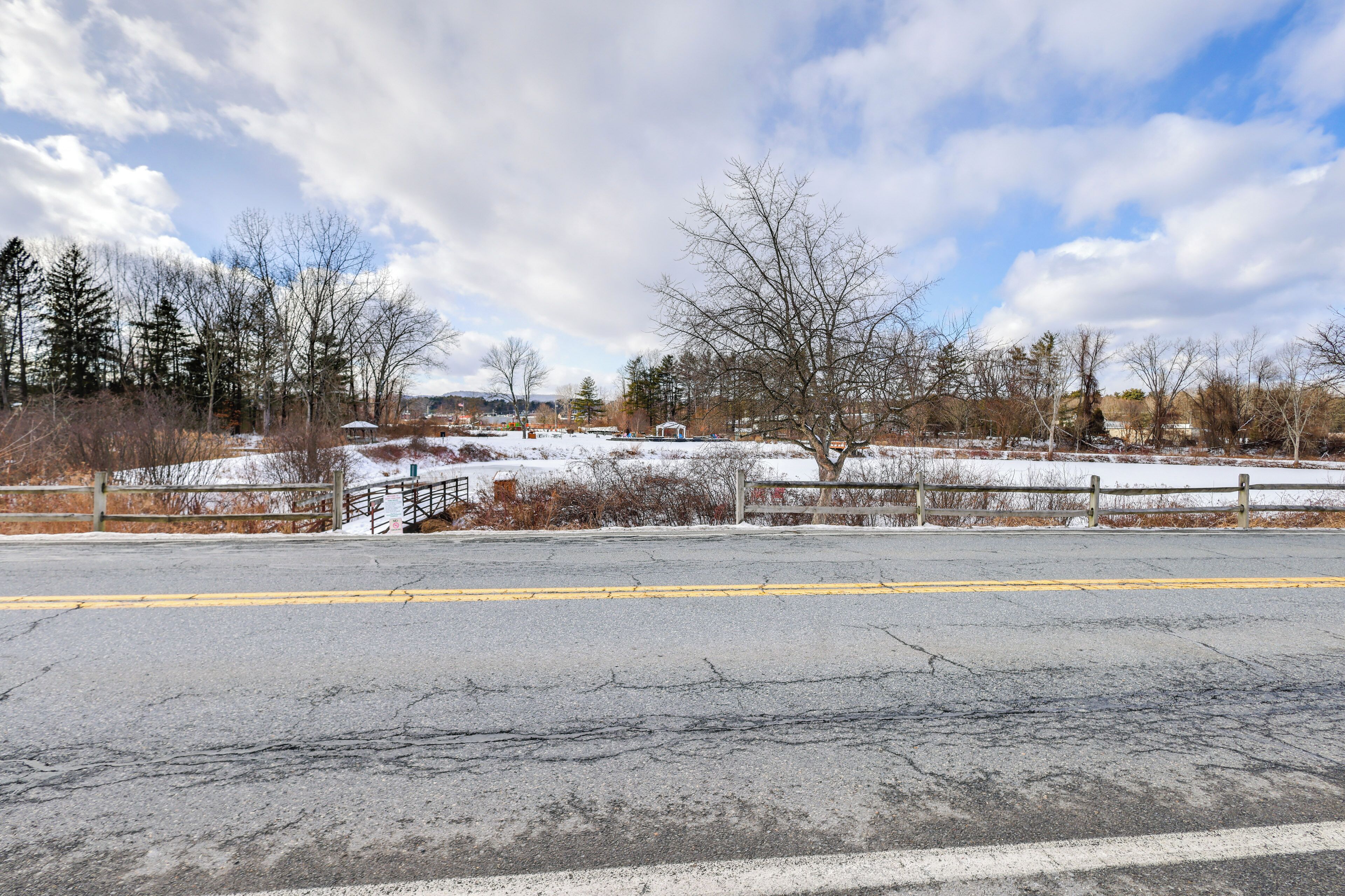 A paved road with a yellow dividing line, a wooden fence, and leafless trees in a rural setting.