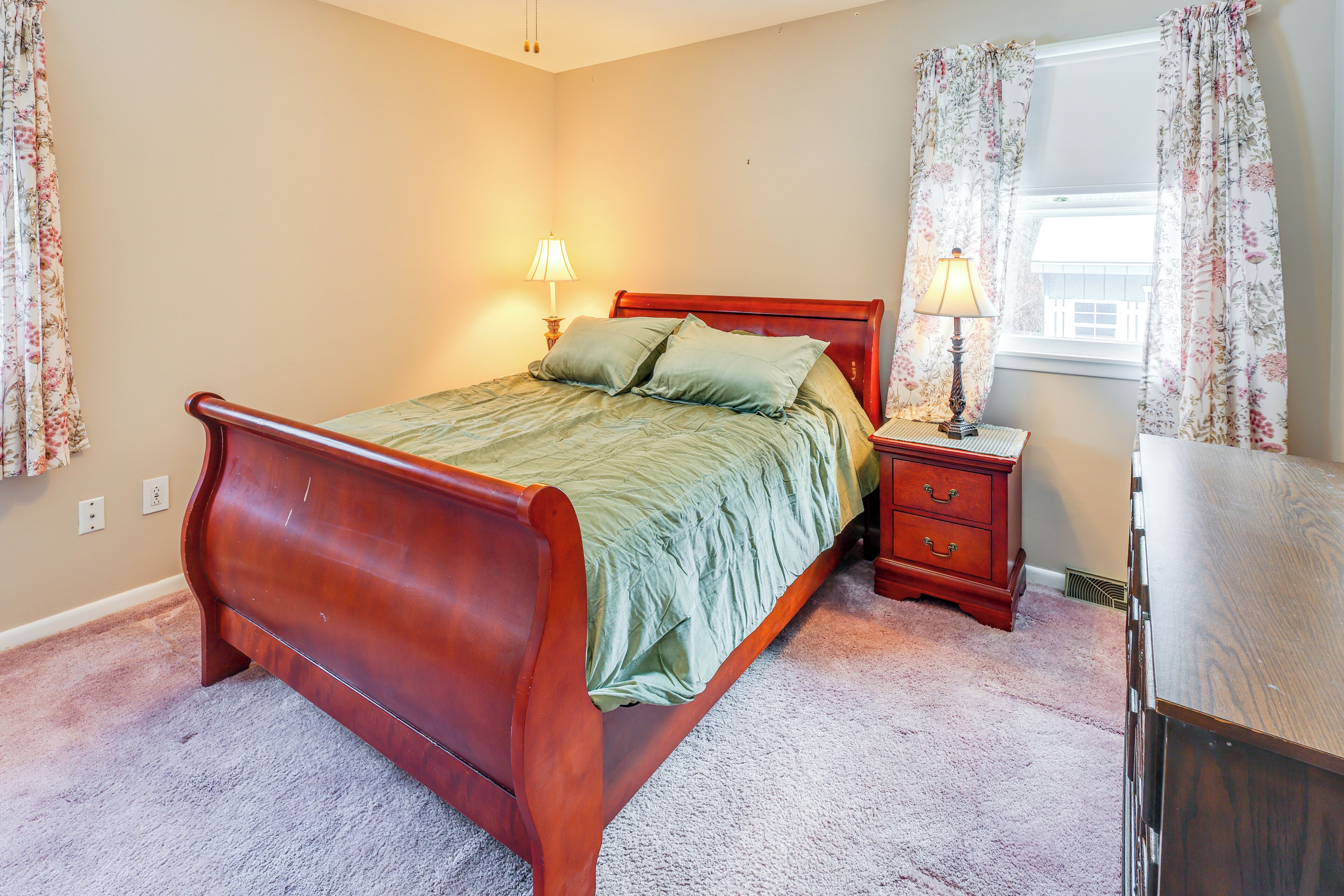 A bedroom with a wooden bed, two bedside tables with lamps, a dresser, and floral curtains.