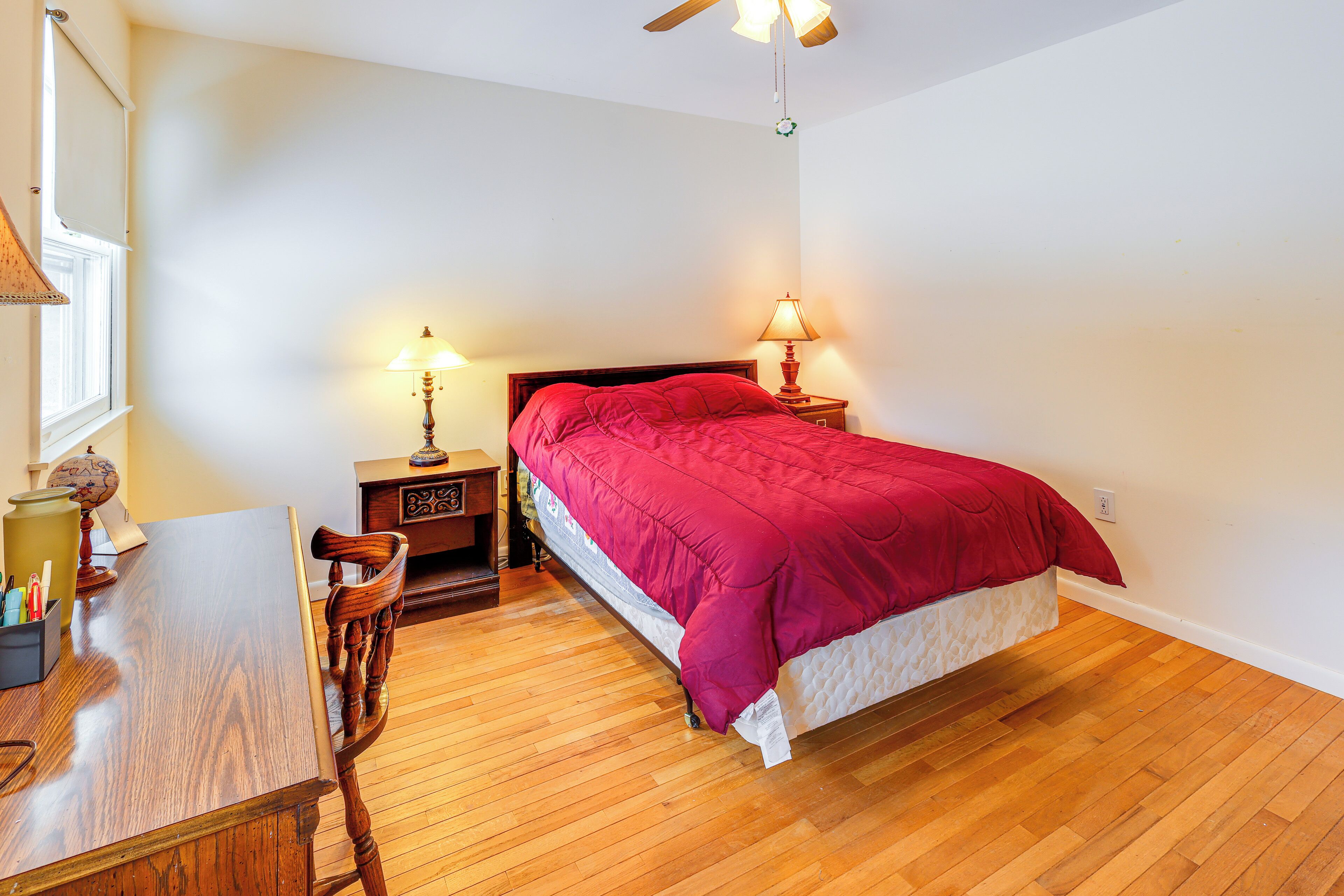 Une chambre avec un plancher en bois, un lit recouvert d’une couverture rouge, deux lampes de chevet, une table de chevet en bois, un bureau avec une chaise et un globe posé sur la table de chevet.