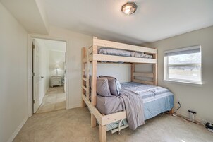 Una cama litera con estructura de madera y una cama individual debajo. Se observa una ventana a la derecha y una puerta que conduce a otra habitación.