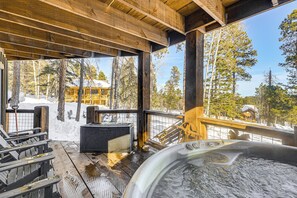 Une terrasse en bois avec un jacuzzi, des sièges d’extérieur et une vue sur un paysage enneigé, avec des arbres et un bâtiment en arrière-plan.