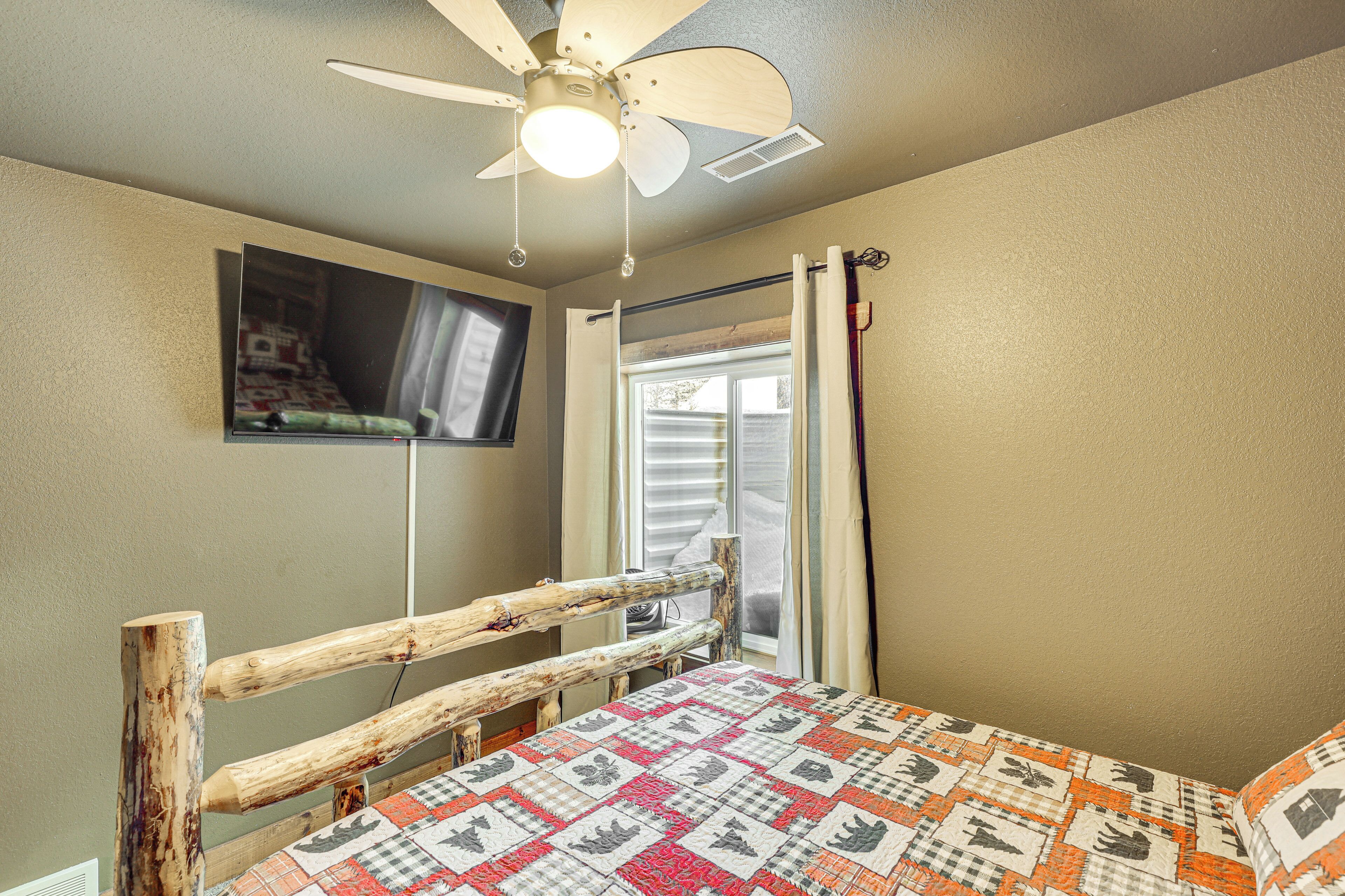 A bedroom with a wooden bed frame, a quilt, a ceiling fan, a television mounted on the wall, and a window with curtains.