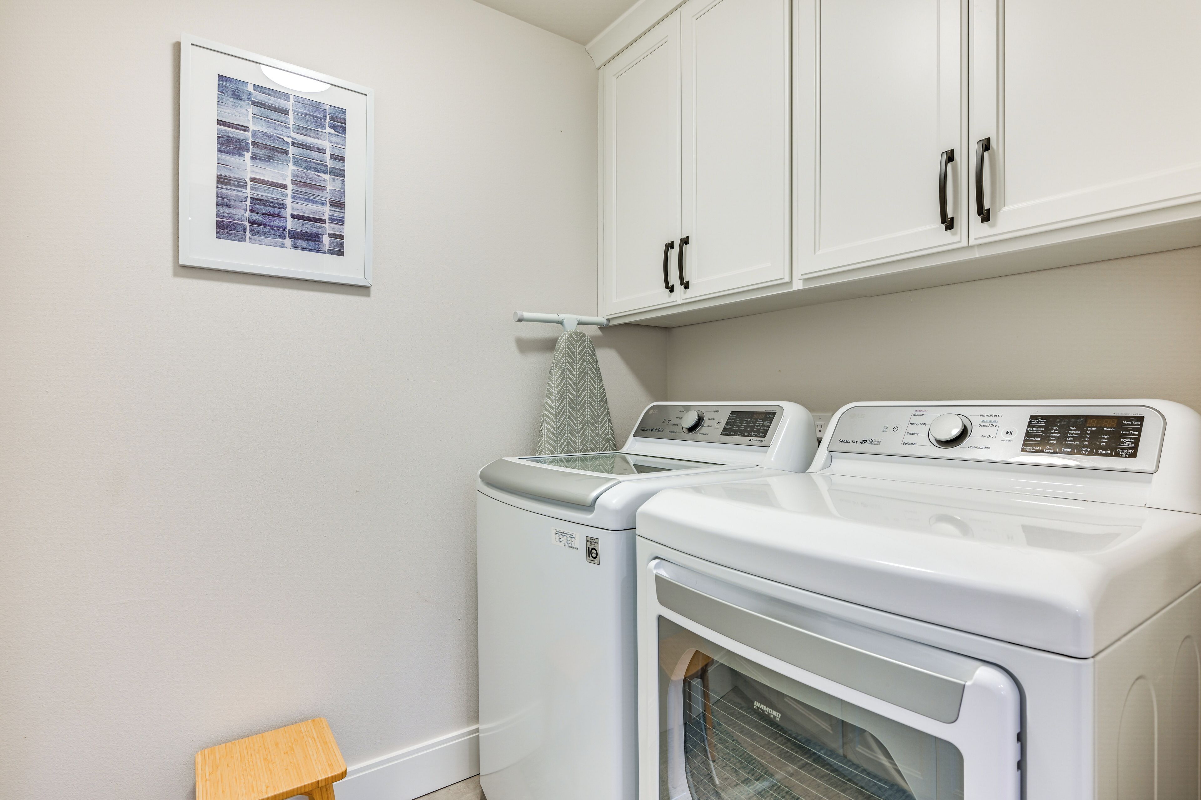 A laundry room with two washing machines and a dryer, white cabinets above, and a framed picture on the wall.