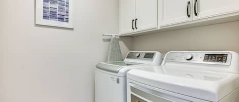 A laundry room with two washing machines and a dryer, white cabinets above, and a framed picture on the wall.