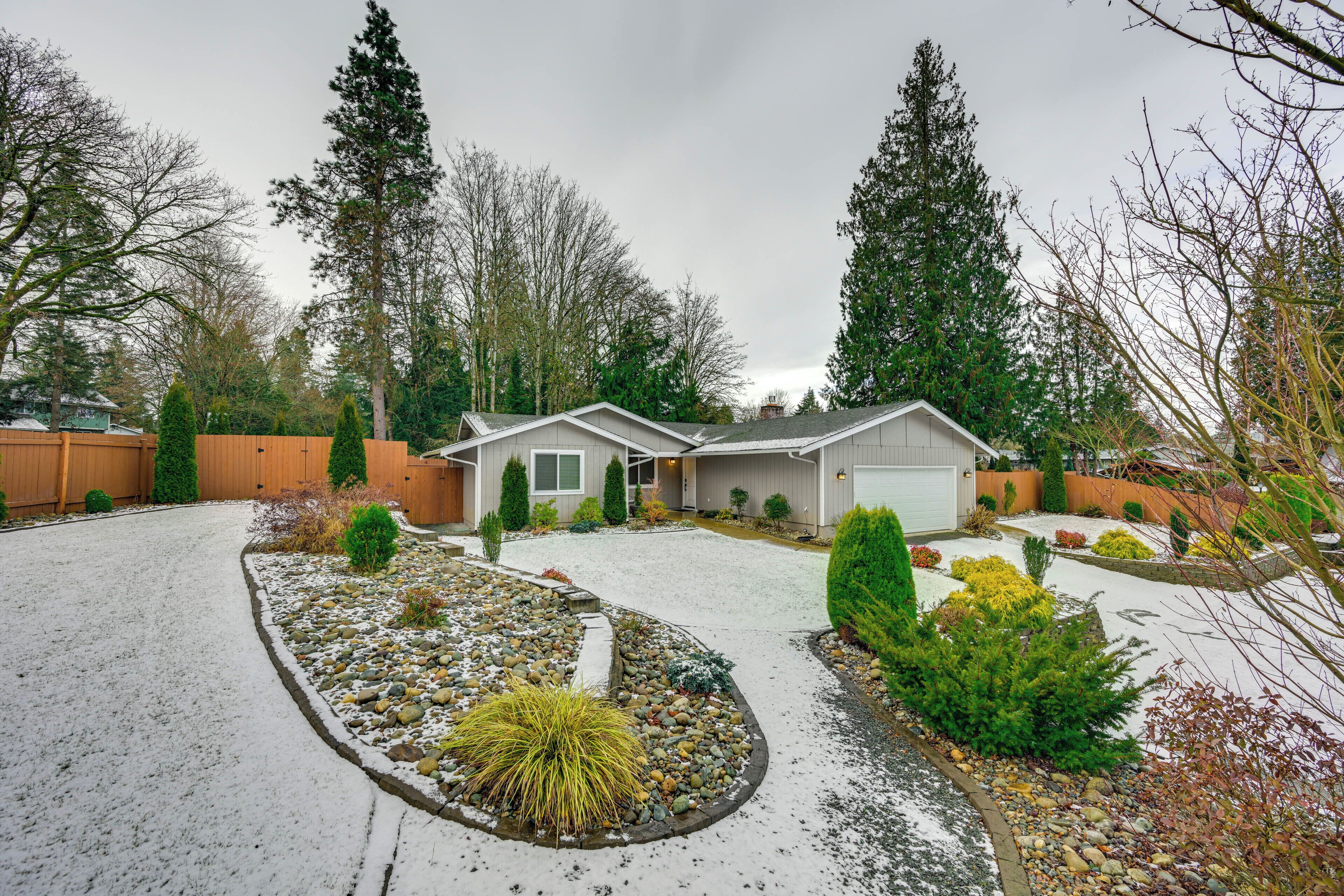 A two-story house with a garage, surrounded by a snow-covered garden with various plants and a gravel driveway.