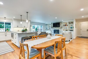 A modern kitchen with white cabinets, a dining area with a wooden table and chairs, and a living area with a fireplace and shelves.