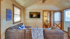 L’intérieur chaleureux d’un chalet avec un plafond en bois, un grand canapé recouvert d’une couverture et un téléviseur fixé au mur.