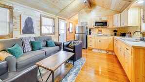 A cozy cabin interior with a wooden ceiling, a grey sofa with teal cushions, a black armchair, a wooden dining table, and a kitchen area with wooden cabinets and appliances. - La Grange Cabin on a Racehorse Farm w/ Pond Views! (La Grange)