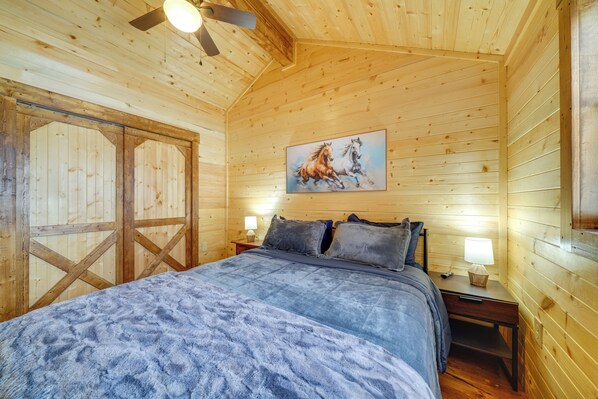 A wooden cabin bedroom with a bed, two nightstands, a ceiling fan, and a painting of horses. - 28 Mi to Downtown Louisville: Serene Farm Getaway (La Grange)