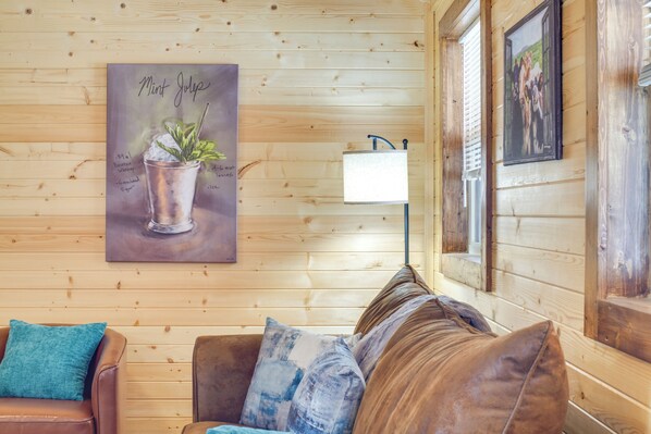 A wooden cabin interior with a brown leather couch, a teal throw pillow, a wall-mounted lamp, and a framed picture of a mint julep. - Horse Farm Retreat w/ Pond View Near Louisville (La Grange)