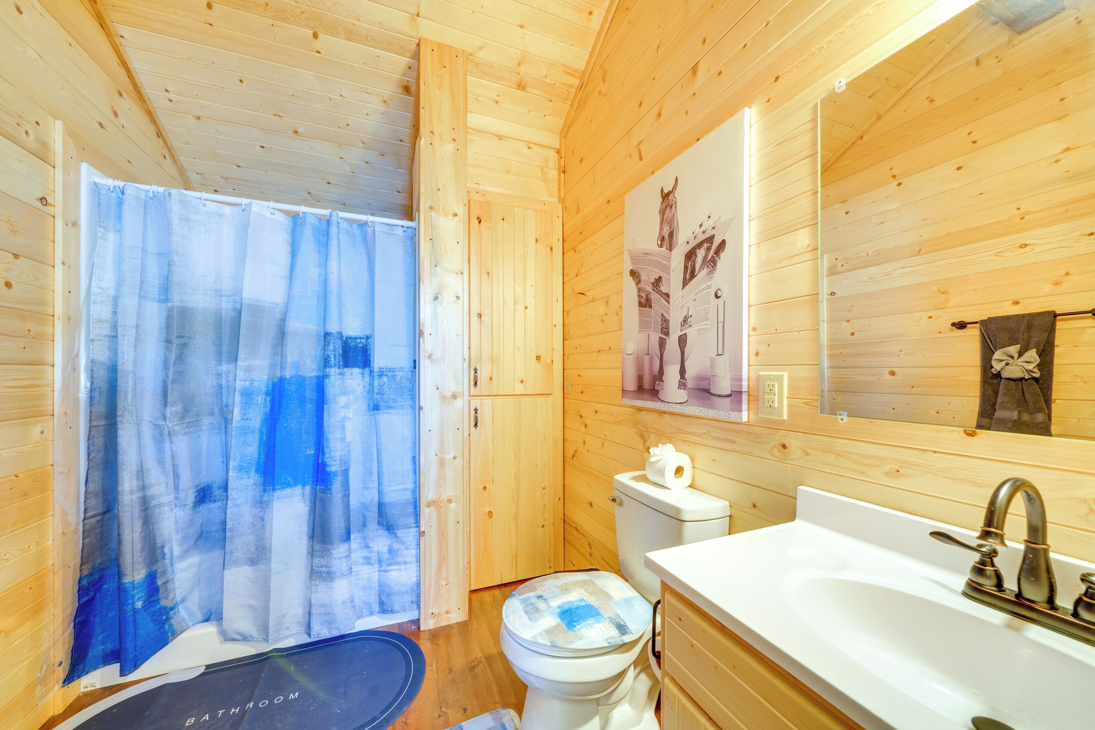 A wooden cabin bathroom with a shower curtain, toilet, sink, and a mirror with a horse-themed decoration.