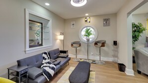 A modern living room with a grey sofa, a bench, a round table with chairs, a large window, and a ceiling light. - < 2 Mi to Ecu: Cozy Greenville Getaway w/ Porch! (Greenville)