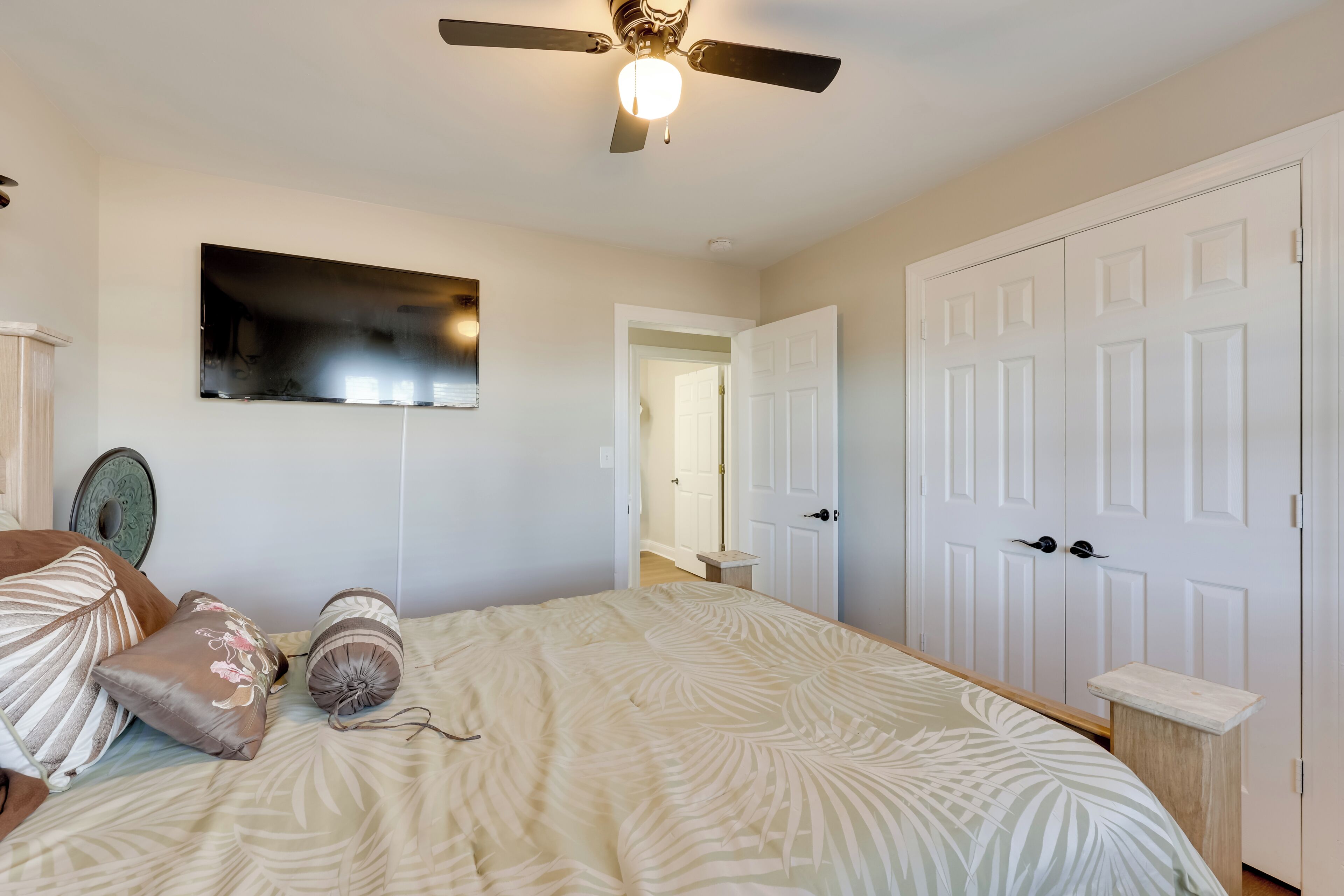 A bedroom with a bed, pillows, a ceiling fan, a wall-mounted TV, and a white door.