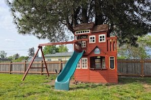 Une maison de jeu rouge avec un toboggan, une balançoire et un arbre en arriÚre-plan.
