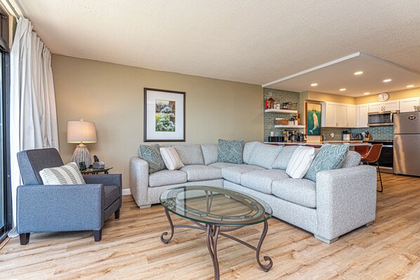 A living room with a sectional sofa, a glass coffee table, and a dining area with a kitchen in the background.