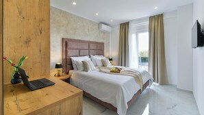 Ein modernes Hotelzimmer mit Schreibtisch aus Holz, einem Bett mit weißen Leinentüchern, einem Wand-Fernseher und einem großen Fenster mit Vorhängen.