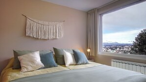 A bedroom with a bed, pillows, a macrame wall hanging, a nightstand with a lamp, and a window with a view of a city and mountains. - Spacious Central Apartment with Garage (San Carlos de Bariloche)