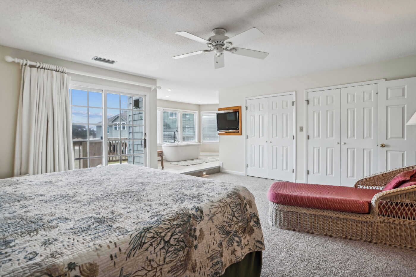 A bedroom with a large bed, a wicker chair, a TV, and a sliding glass door to the balcony.