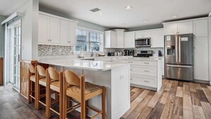 Uma cozinha moderna com armários brancos, uma ilha grande com banquetas, eletrodomésticos de aço inoxidável e piso de madeira em padrão herringbone.