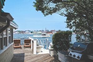 Une terrasse avec des chaises en bois, un barbecue et une vue sur l’eau, avec des bateaux et des bâtiments en arrière-plan.
