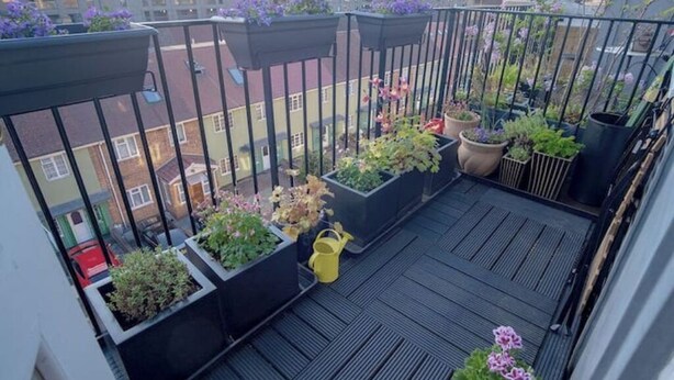 A balcony with multiple planters containing various flowers and plants, a yellow watering can, and a view of surrounding buildings. - Airy one-bed in Limehouse with views of the city (London)