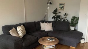 A living room with a dark grey sectional sofa, a round wicker coffee table, and a floor lamp. There are several potted plants and a framed picture on the wall. - Airy one-bed in Limehouse with views of the city (London)