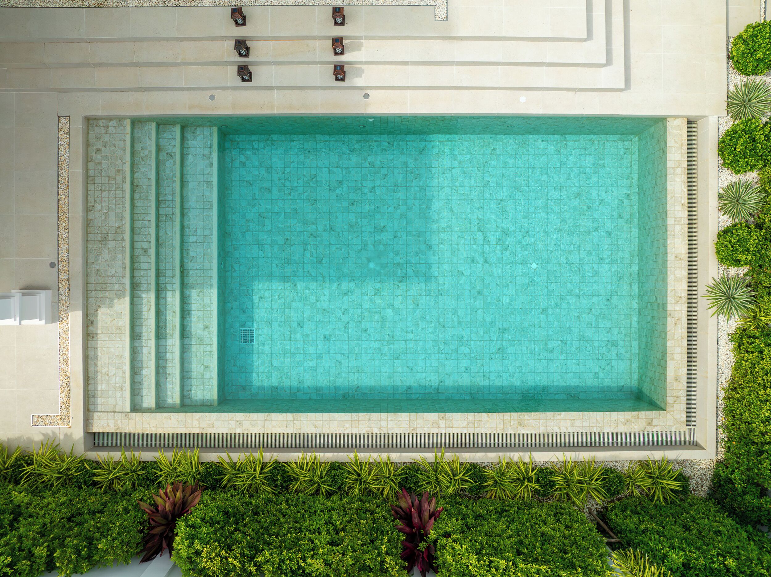 A rectangular swimming pool with a tiled edge, surrounded by greenery and a tiled walkway.