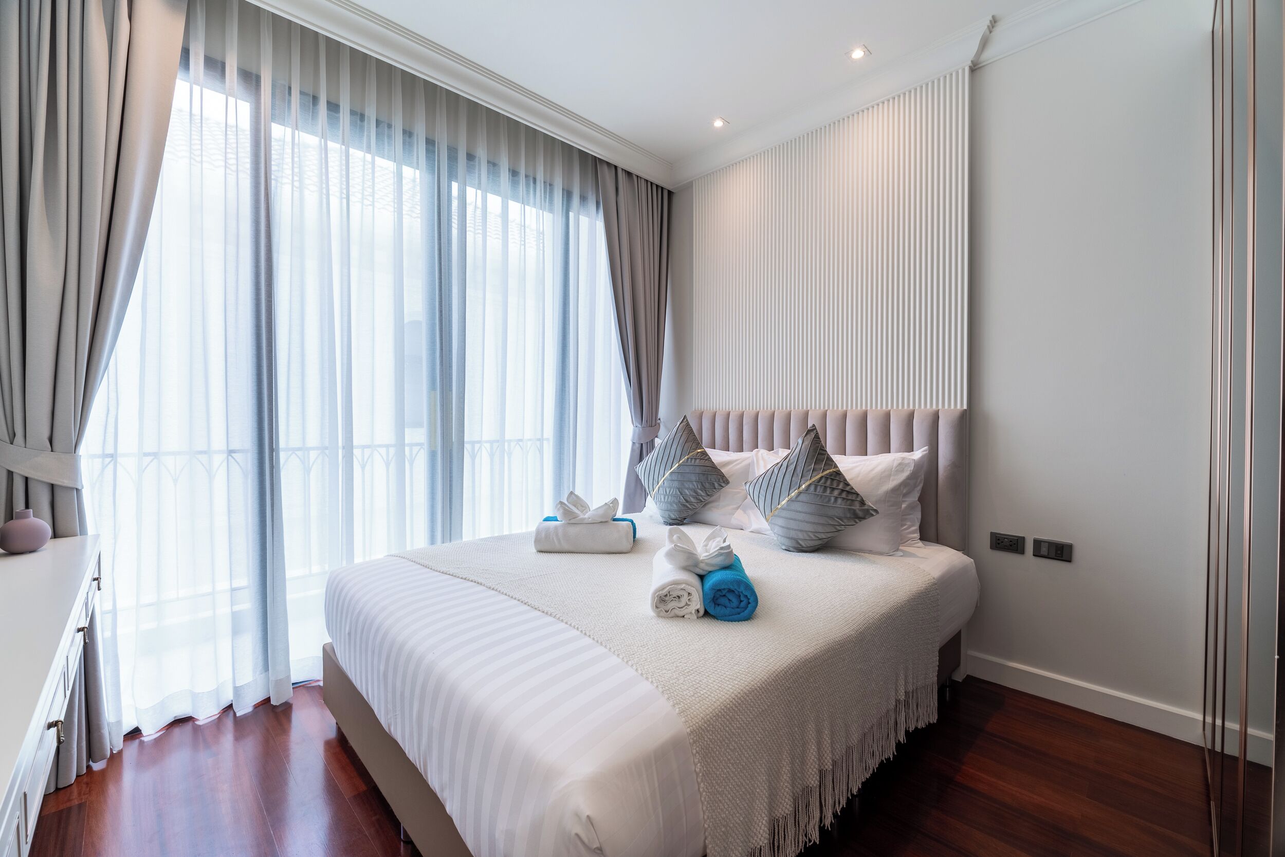A neatly made bed with a white bedspread and pillows, a bedside table with a lamp, and a large window with sheer curtains.