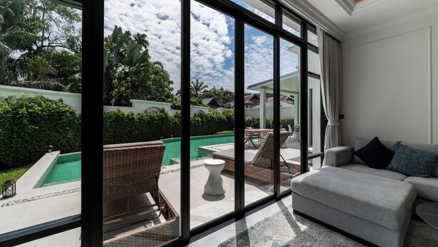 A spacious living area with a large window overlooking a swimming pool and lush greenery. - FIFTH ELEMENT pool villa by NLA (Choeng Thale)
