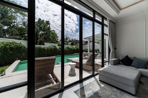 A spacious living area with a large window overlooking a swimming pool and lush greenery. - FIFTH ELEMENT pool villa by NLA (Choeng Thale)
