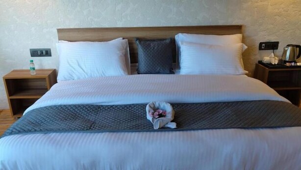 A neatly made bed with white and gray linens, a wooden headboard, and a small bedside table with a water bottle and a power outlet. - Anupraas Resort (Dewalkhal)