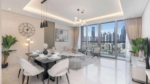A modern living room with a dining area, large windows with a city view, and a large wall clock. - StayShort - 3 Bedroom Apartment in Sterling West with Full Burj Khalifa Views (Business Bay)