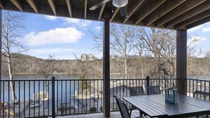 Um deck coberto com mesa e cadeiras, com vista para um lago e edifícios ao fundo.