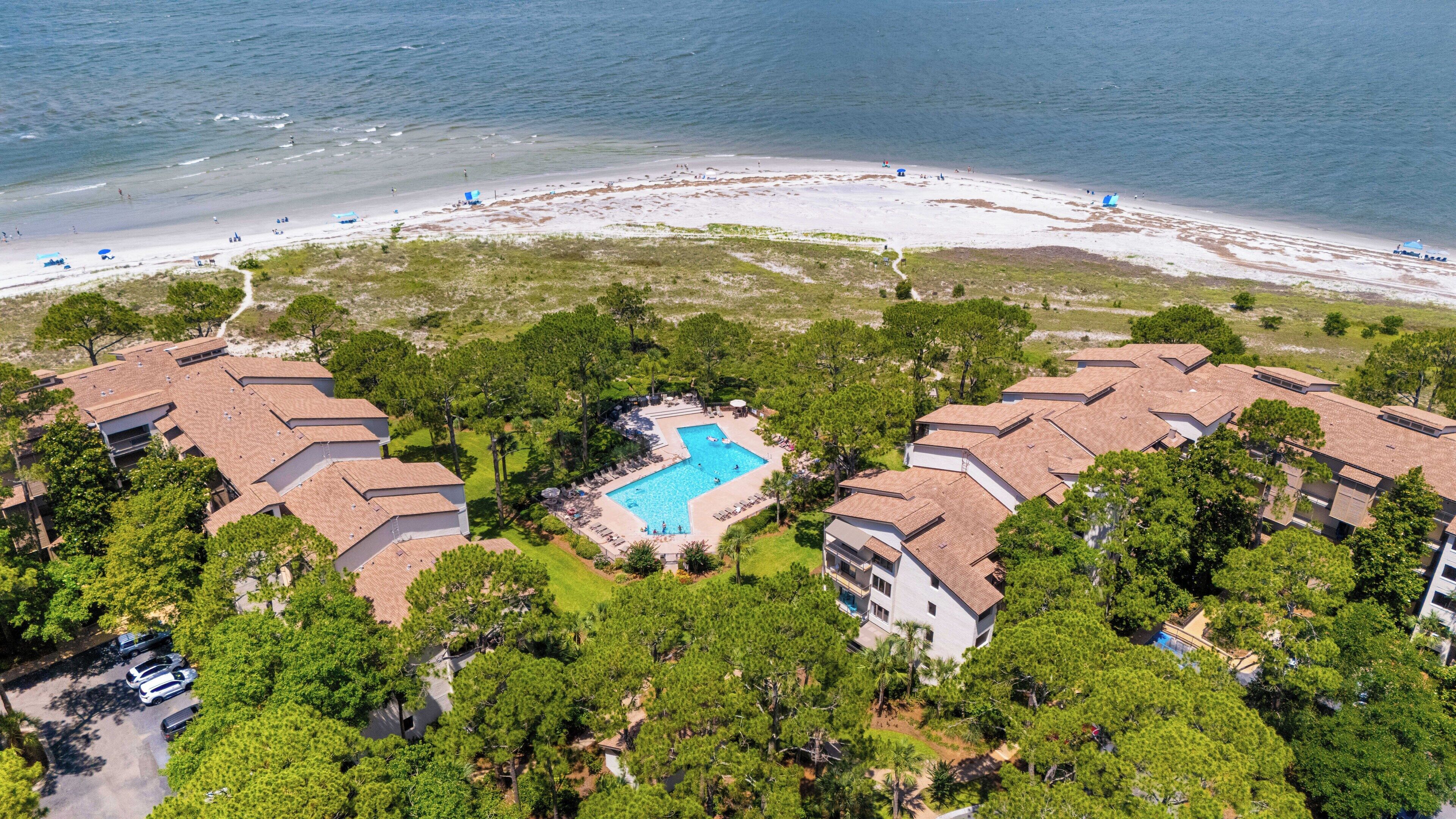 An aerial view of a resort with a swimming pool surrounded by lush greenery, a beach, and multiple buildings with a consistent architectural style.