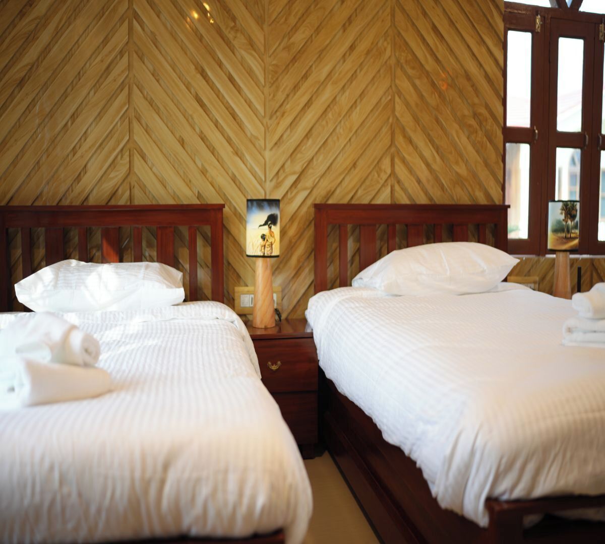 A hotel room with two beds, wooden headboards, a wooden herringbone-patterned wall, a nightstand with a lamp, and a painting.