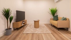Ein modernes Wohnzimmer mit einem Flachbildfernseher, einem Holzschrank, einem runden Couchtisch, einem beigen Sofa und zwei Topfpflanzen.