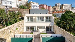 Un edificio moderno de dos pisos con grandes ventanales y techo plano, rodeado por un muro de piedra y vegetación.
