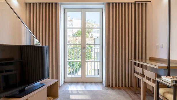 Una sala de estar moderna con una televisión de pantalla plana sobre un soporte de madera, una mesa de centro de vidrio, un sofá y una puerta corrediza de vidrio que da a un balcón con vistas a edificios y follaje.
