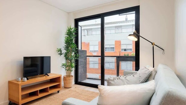 A modern living room with a flat-screen TV on a wooden stand, a grey sofa, a floor lamp, a potted plant, and a large window with a view of buildings.