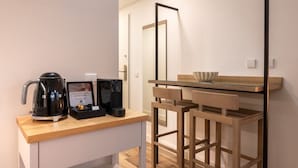 Une petite cuisine moderne équipée d’une machine à café, d’une bouilloire et d’un petit réfrigérateur. On y trouve également un comptoir-bar en bois sur lequel est posé un bol.