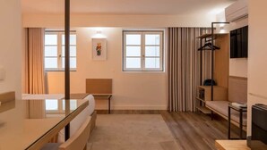 Une chambre d’hôtel moderne dotée d’une cloison en verre, d’un banc en bois, d’un canapé, d’une télévision et d’un porte-manteau suspendu.