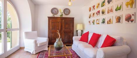 Un salón con un sofá blanco, cojines rojos, un aparador de madera y una pared decorada con pinturas de frutas y verduras.