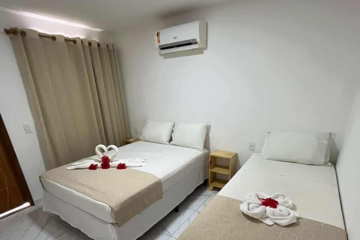 A hotel room with two beds, a small wooden nightstand, a wall-mounted air conditioner, and a window with curtains.