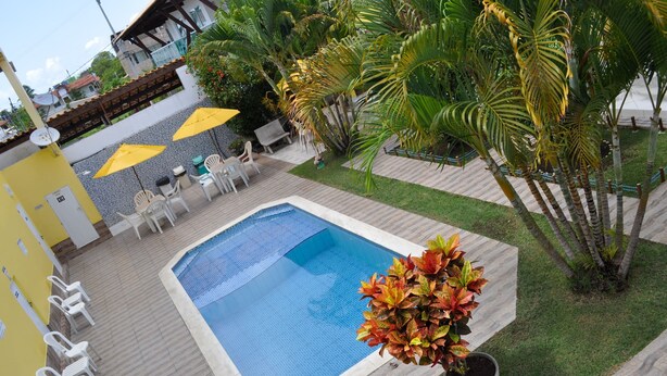 A swimming pool with a tiled deck, surrounded by palm trees and a yellow building. - Pousada e Flat Diamant's (Ipojuca)