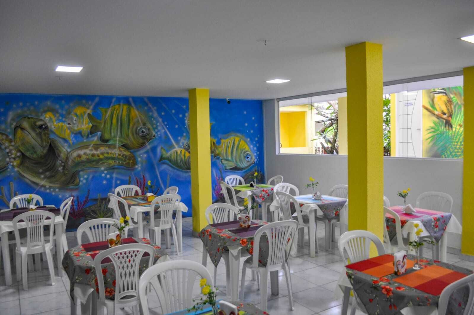 Restaurante com cadeiras e mesas de plástico branco, um mural de peixes na parede e pilares amarelos.