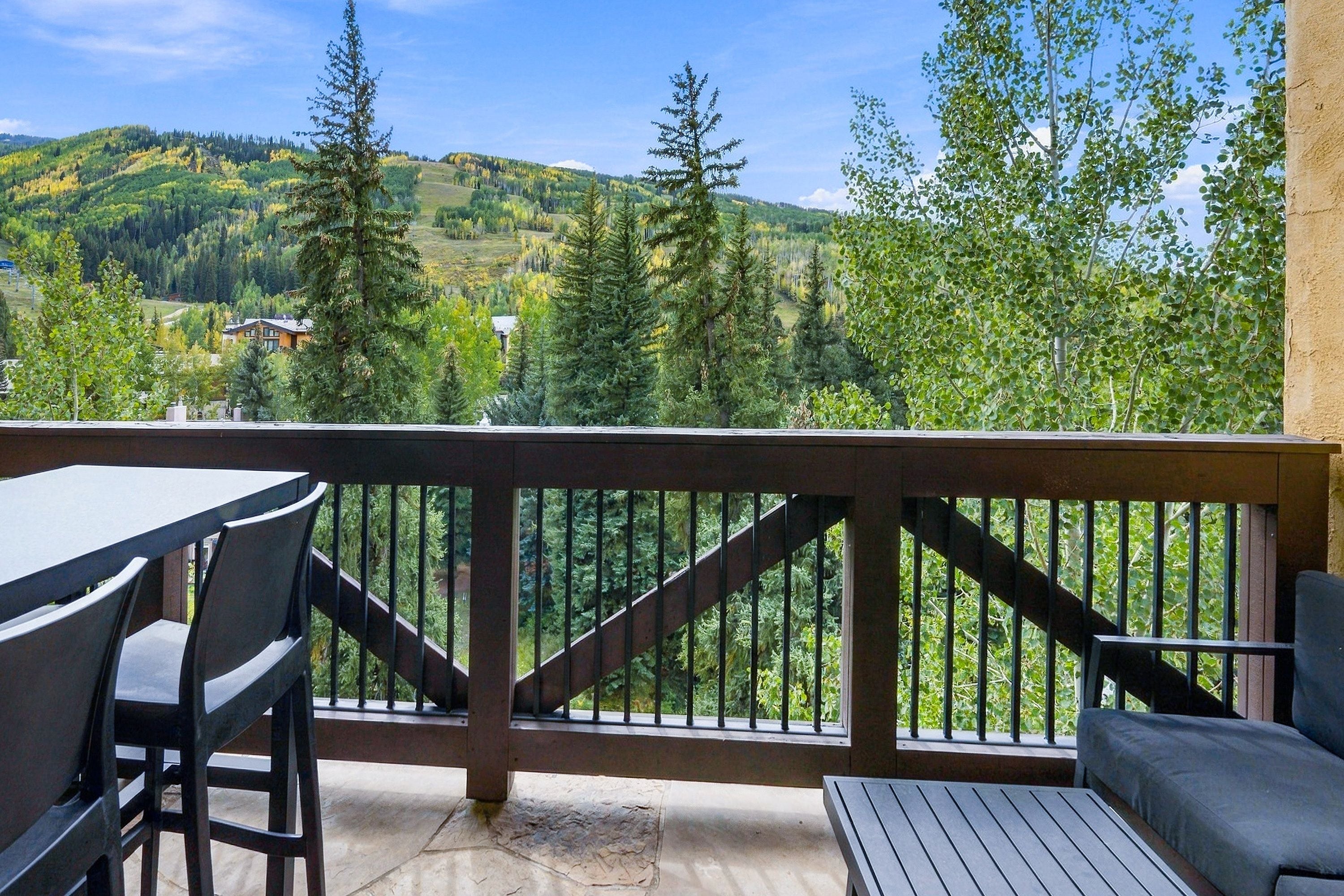 Un balcon avec une table et des chaises, donnant sur un paysage montagneux parsemé d’arbres et de bâtiments.
