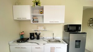Una cocina compacta con armarios blancos, microondas integrado, refrigerador, lavamanos y cafetera.