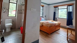 A room with a wooden bed, a window with curtains, a bathroom with a toilet and shower, and a view of greenery outside. - Bon Kiputhang (Chungthang)