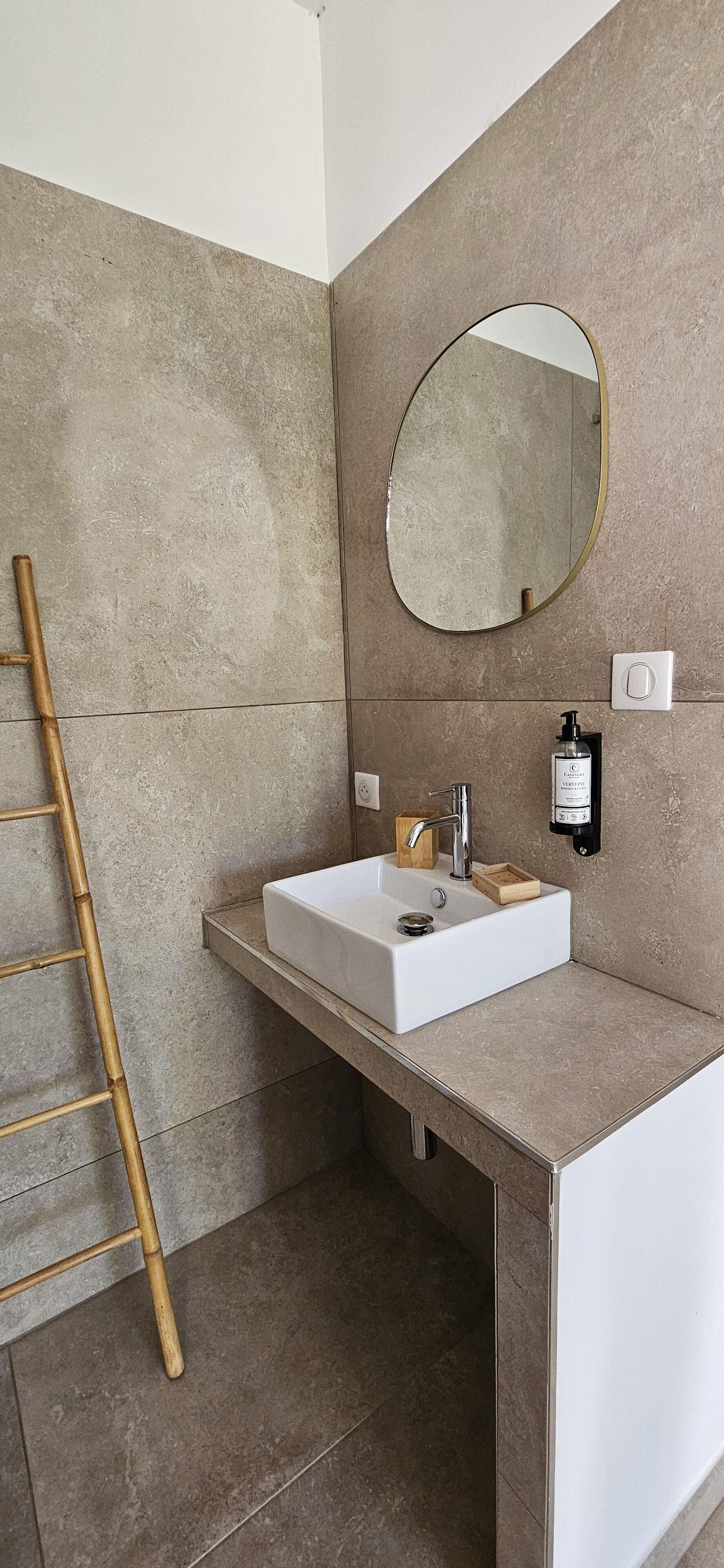 Une salle de bain avec un lavabo blanc, un miroir rond et un escabeau en bambou.